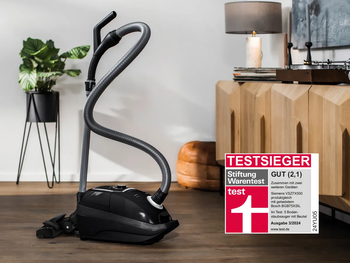 Ein schwarzer Siemens Beutelstaubsauger steht auf einem dunklen Boden in einem modernen Raum, umgeben von einem Holz-Sideboard und einer grünen Topfpflanze vor einer weißen Wand.