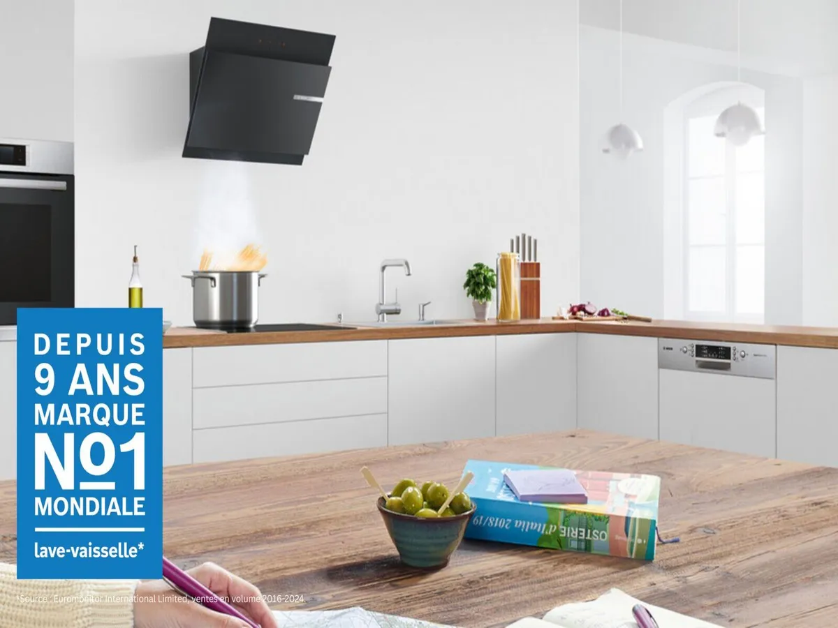 Cuisine moderne avec une hotte noire élégante, une casserole fumante sur le feu et une table en bois avec un bol d'olives et des magazines colorés.