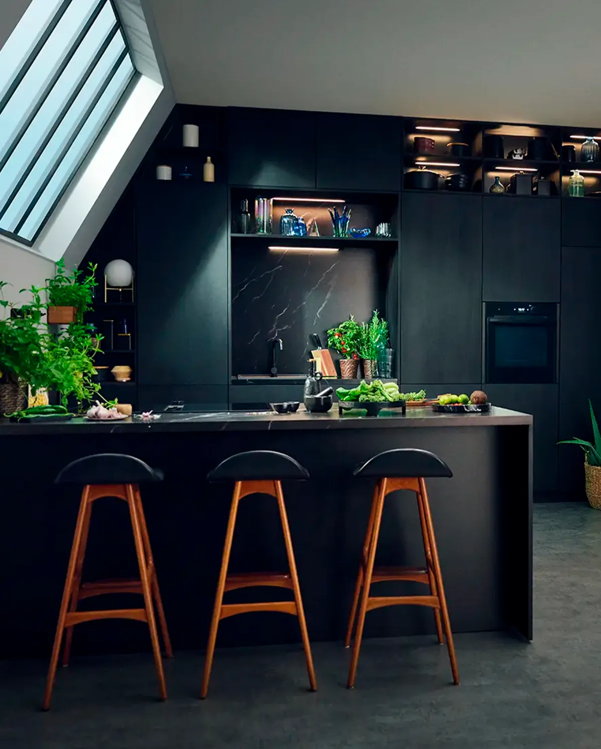 „O bucătărie cu suprafețe închise la culoare și o insulă de bucătărie înaltă, iar în spate se află un cuptor încorporat.