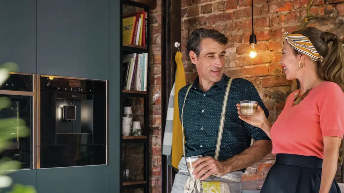 A man and woman stand together in a modern kitchen, smiling and facing the camera. 