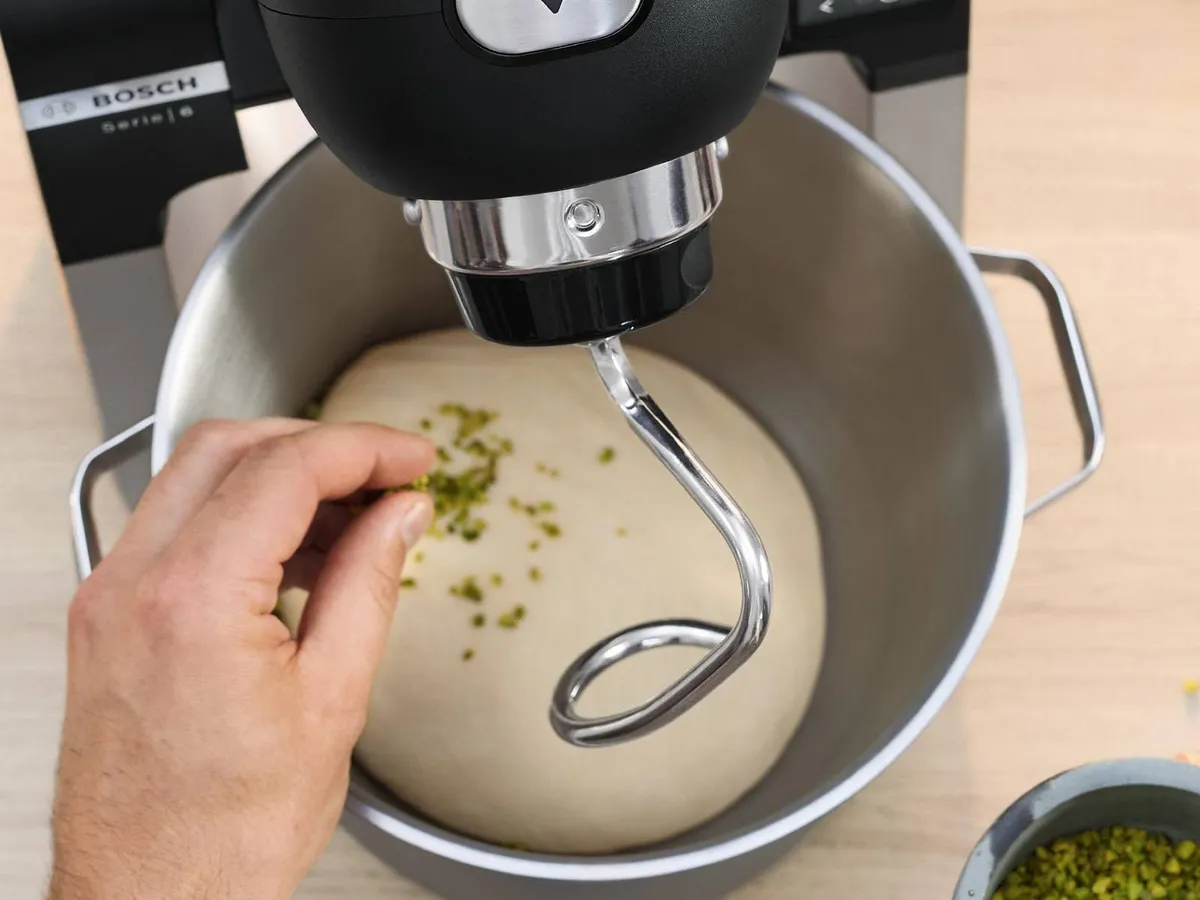 Een hand strooit gehakte pistachenoten over het deeg in een Bosch-keukenmixer, omringd door eieren en bakmaterialen op een houten oppervlak.