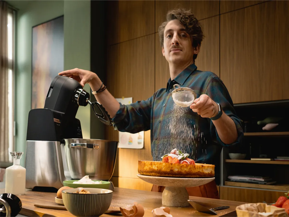 Une personne prépare un dessert dans une cuisine chaleureuse, en saupoudrant du sucre glace sur un cheesecake à la fraise posé sur un support.