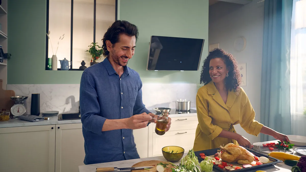 A man and woman prepare food together in a kitchen with an angled cooker hood above the stove.  
