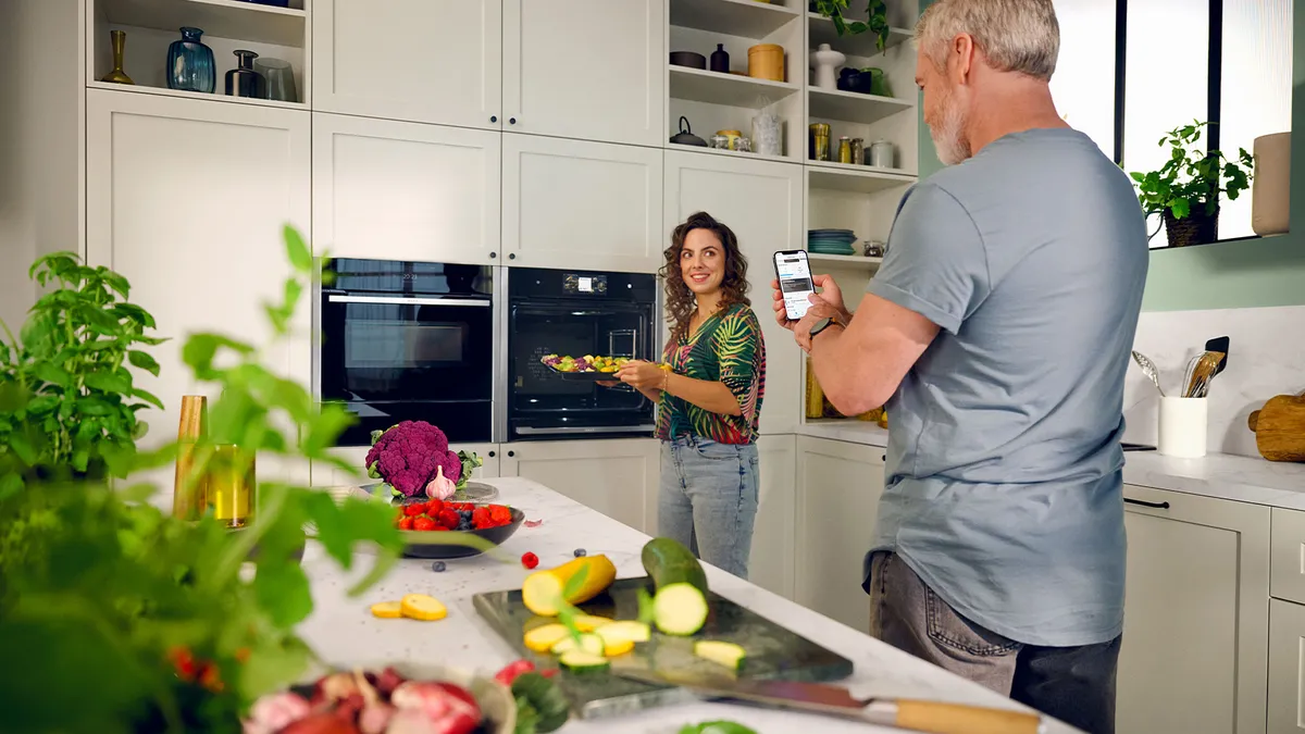 A man and woman are engaged in food preparation in a kitchen featuring an angled cooker hood above the cooking area.  