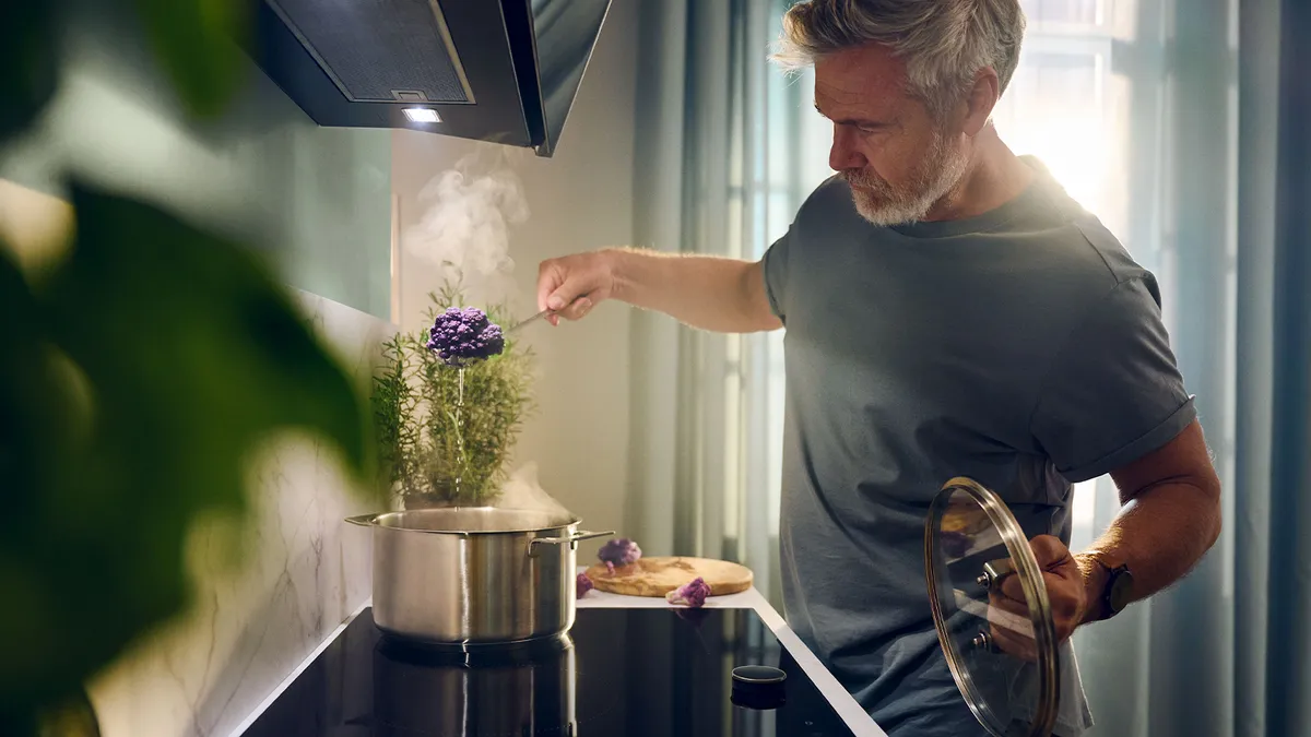 A man cooks in a kitchen, stirring a pot filled with fresh herbs on the stove.