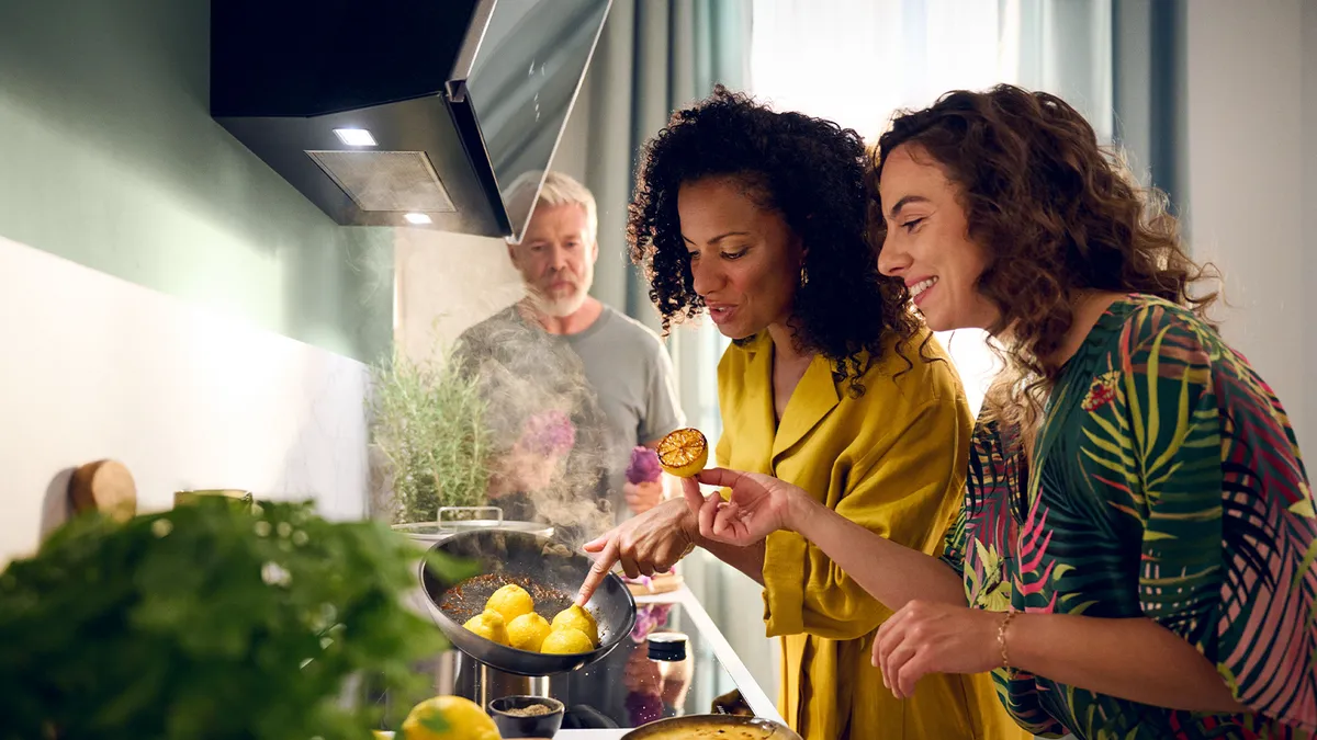 Two women cook together in a kitchen, while a man stands in the background observing their activity.