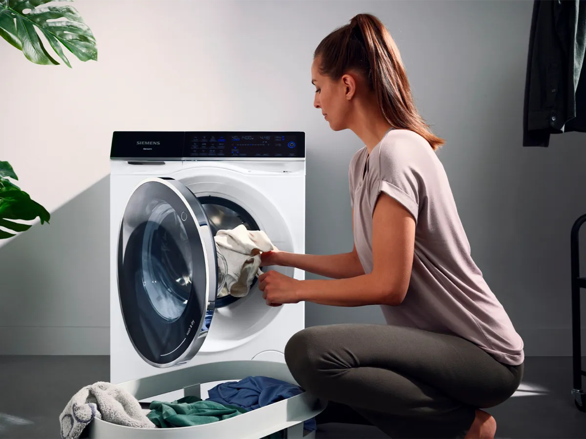 Eine Frau im Freizeitoutfit kniet neben einer modernen Waschmaschine und belädt sie mit heller Wäsche aus einem daneben stehenden Korb.
