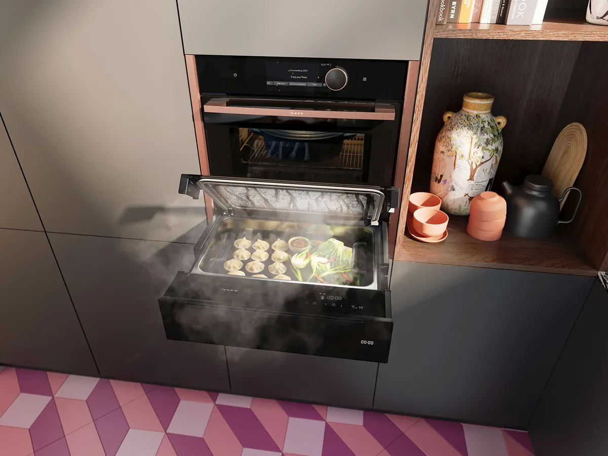 Съвременна кухненска сцена с лапа за пара, пълна с пелмени и зеленчуци, до декоративна керамика и книги.