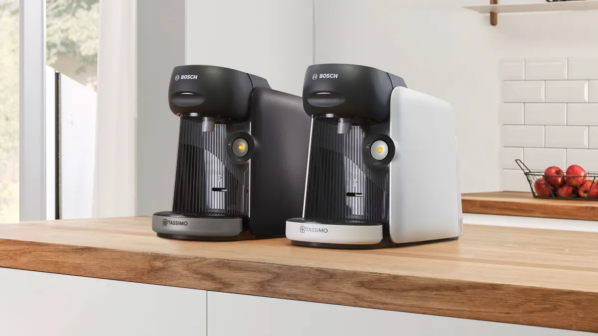 Two Bosch Tassimo coffee machines in black and white sit on a wooden kitchen countertop, against a bright, modern background.