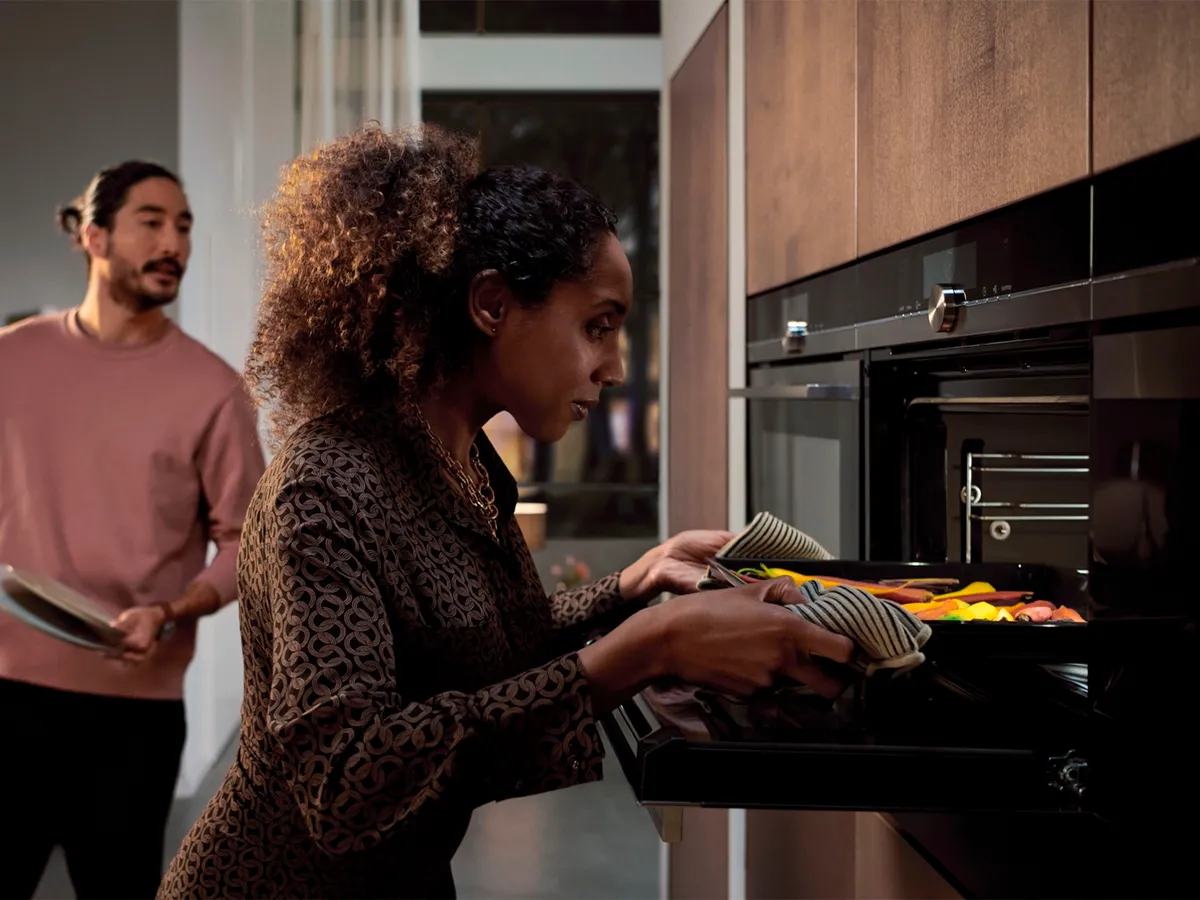 En kvinde tager en bageplade med grøntsager ud af en ovn, mens en mand står i baggrunden med en tallerken i hånden.