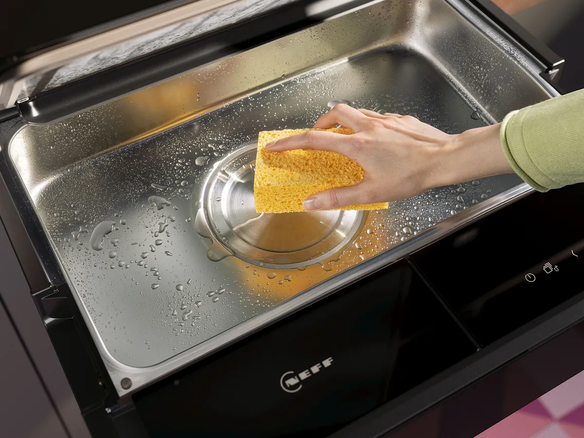 Eine Hand hält einen gelben Schwamm über einer nassen Edelstahlkochfläche, mit sichtbaren Tropfen und Dampf, was darauf hinweist, dass gerade gereinigt wird.