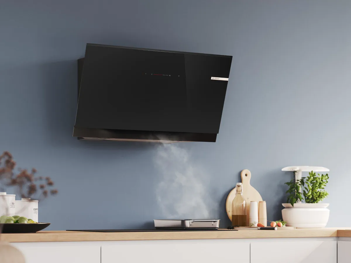 Une hotte de cuisine noire élégante montée sur un mur bleu, avec de la vapeur s'élevant d'une zone de cuisson en dessous, avec des planches à découper et des plantes.