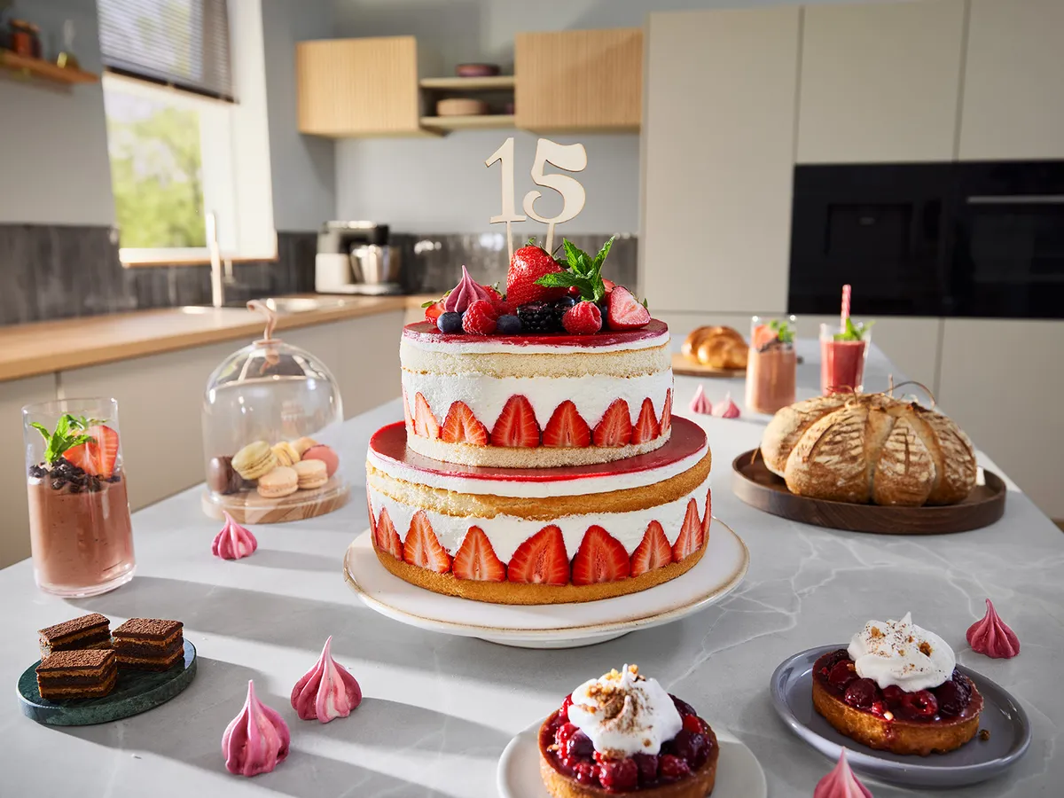 Eine Arbeitsplatte in der Küche mit verschiedenen Backwaren darauf und einer großen Torte in der Mitte, in der Kerzen stecken, die die Zahl 15 anzeigen.