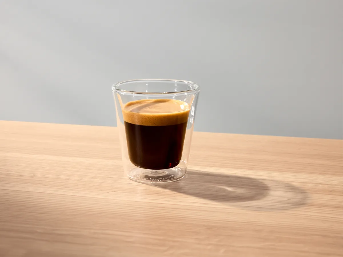 A clear glass cup filled with a rich espresso layered with a crema sits on a wooden surface. Soft light creates a gentle shadow beside the cup, enhancing the warm tones of the coffee.