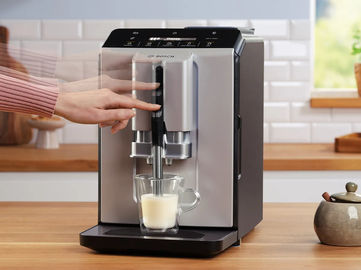 A person is pressing the button on a Bosch coffee machine to brew a drink. A clear glass cup containing milk is positioned under the spout, and the sleek machine features a modern silver and black design. The background shows a bright kitchen with white tile and wooden countertops.
