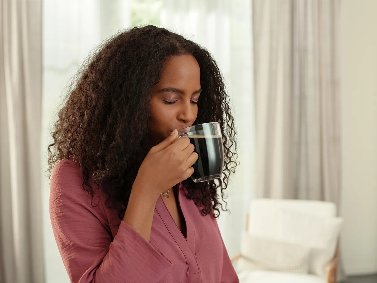 A person with curly hair holds a glass of dark beverage close to their face, enjoying a moment of relaxation. They are dressed in a light purple top, and the background features soft, neutral-colored furnishings and sheer curtains.