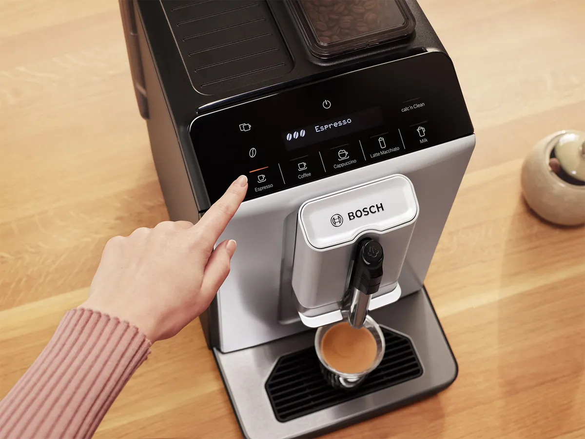 An overhead view of a Bosch coffee machine with a finger pressing the "Espresso" button on the control panel. The machine has a sleek design, and a cup containing freshly brewed espresso sits below the spout on a wooden countertop.