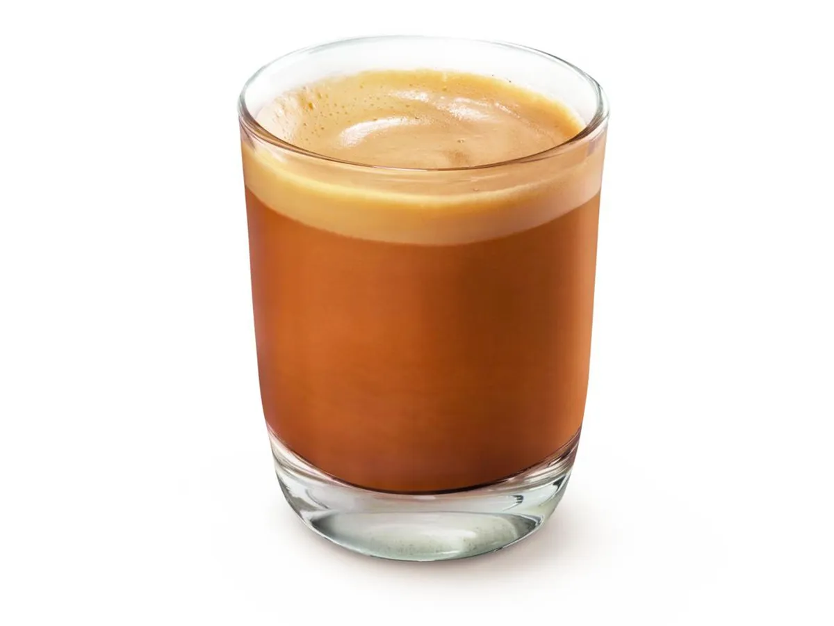 A clear glass filled with rich, dark espresso topped with a creamy layer of froth, sitting on a light surface.