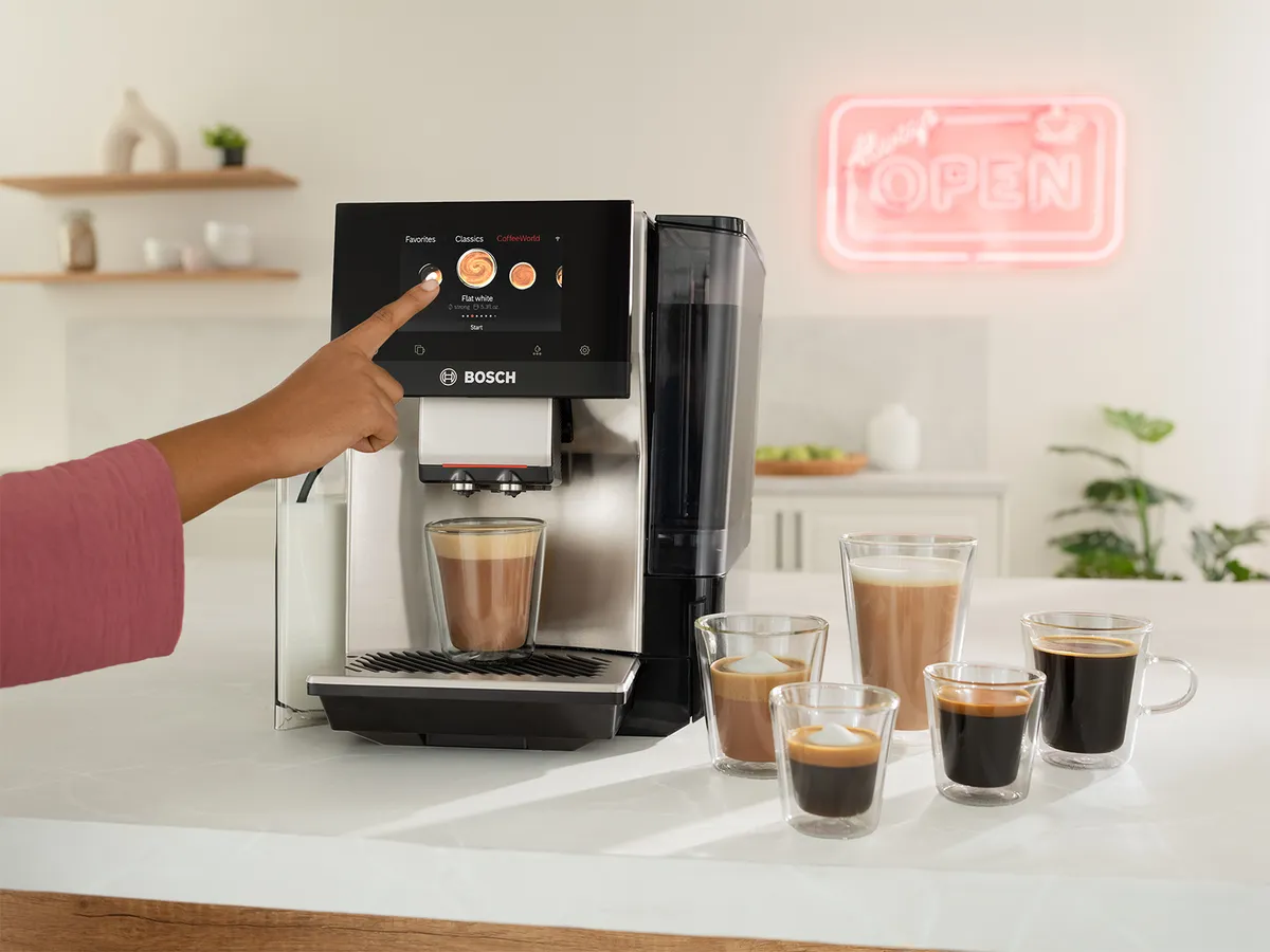 A person in a pink shirt is selecting a drink on a Bosch coffee machine's touch screen. A latte macchiato is being prepared, with cups of cappuccino and espresso displayed in front. A neon "OPEN" sign is visible in the background.