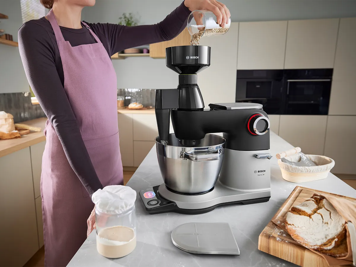 Die Serie 8 Küchenmaschine steht auf einer Küchenarbeitsplatte, umgeben von Brot und Zutaten, während eine Person die Getreidemühle an der Maschine verwendet, um Körner zu mahlen.