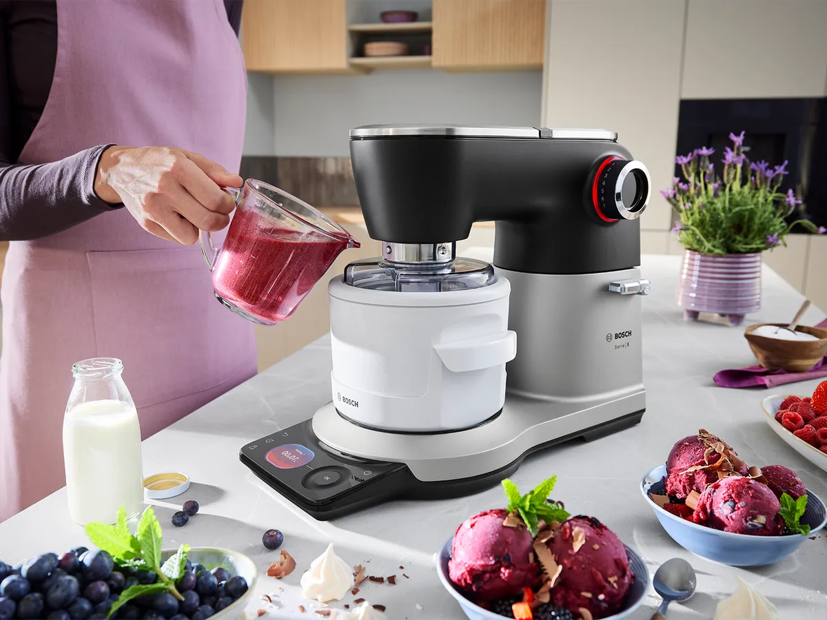 Die Serie 8 Küchenmaschine steht auf einer Küchenarbeitsplatte, umgeben von Eiscreme und Zutaten, während eine Person eine Flüssigkeit in das Eismaschinenzubehör der Maschine gießt, um Eiscreme herzustellen.