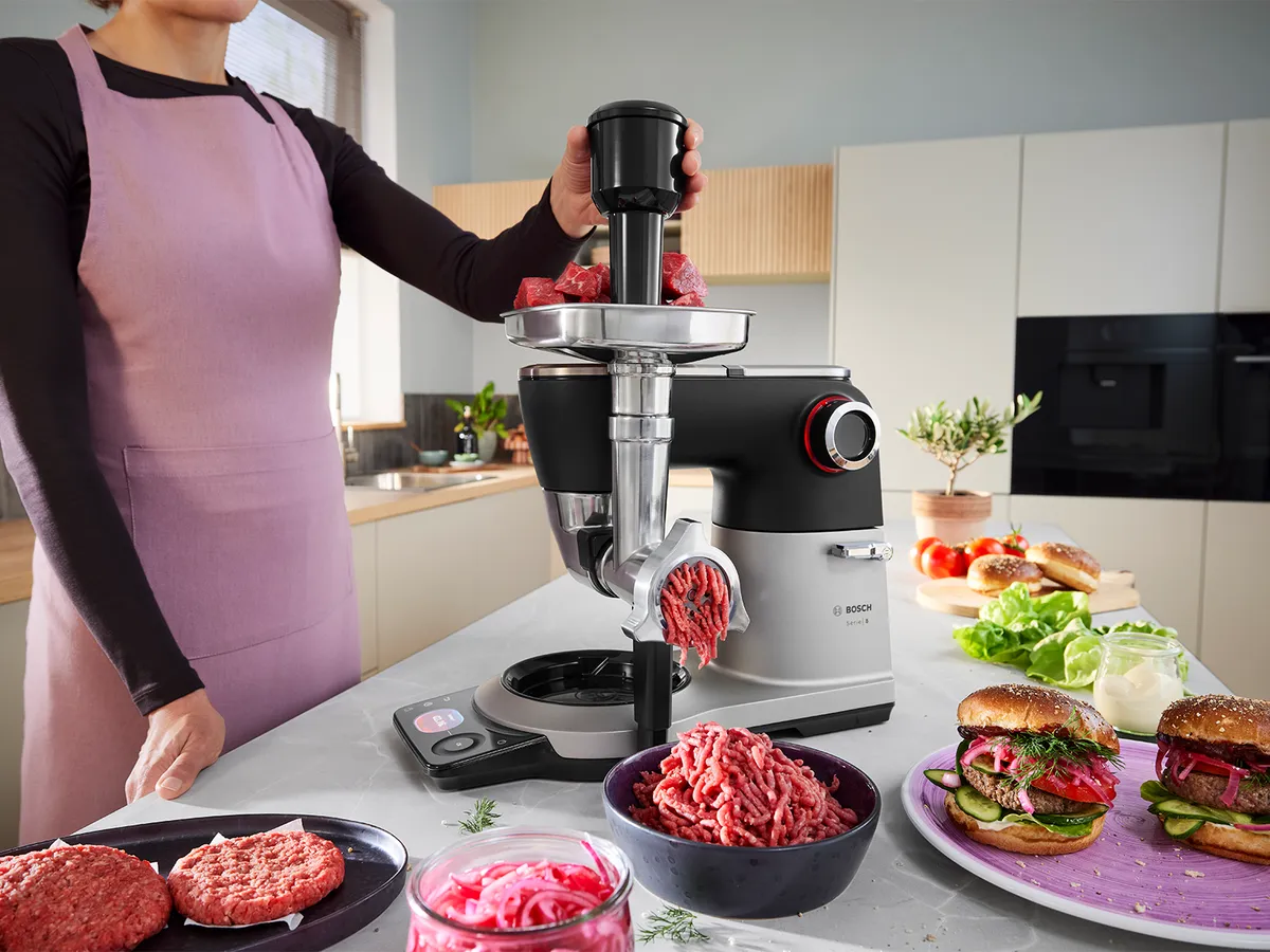 Die Serie 8 Küchenmaschine steht auf einer Küchenarbeitsplatte, umgeben von verschiedenen Fleischgerichten, während eine Person den Fleischwolf an der Maschine verwendet, um Hackfleisch herzustellen.