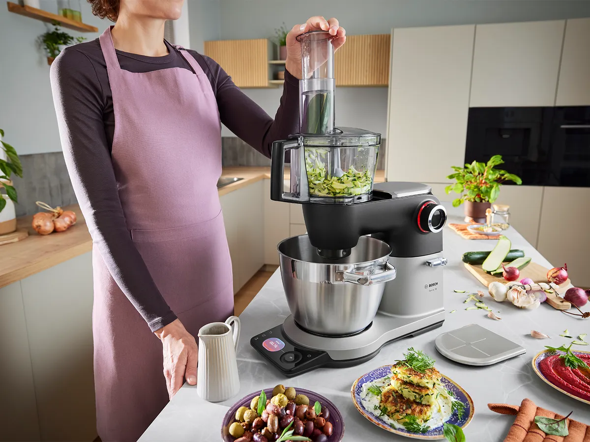 Die Serie 8 Küchenmaschine steht auf einer Küchenarbeitsplatte, umgeben von verschiedenen Gerichten, während eine Person das Multi-Mixer-Zubehör an der Maschine verwendet, um Zutaten zu vermischen.