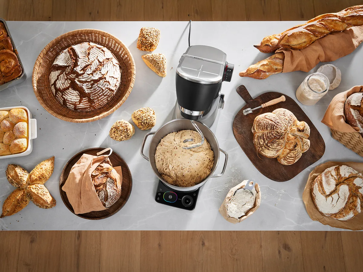 Verschiedene Brotsorten und Gebäck auf einer Marmoroberfläche mit einem schwarzen Standmixer, der Teig knetet.