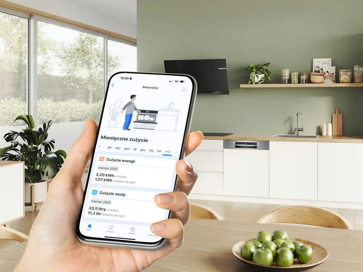 Ręka trzymająca smartfon wyświetlający aplikację zużycia energii w domu. Nowoczesna kuchnia w tle. 
