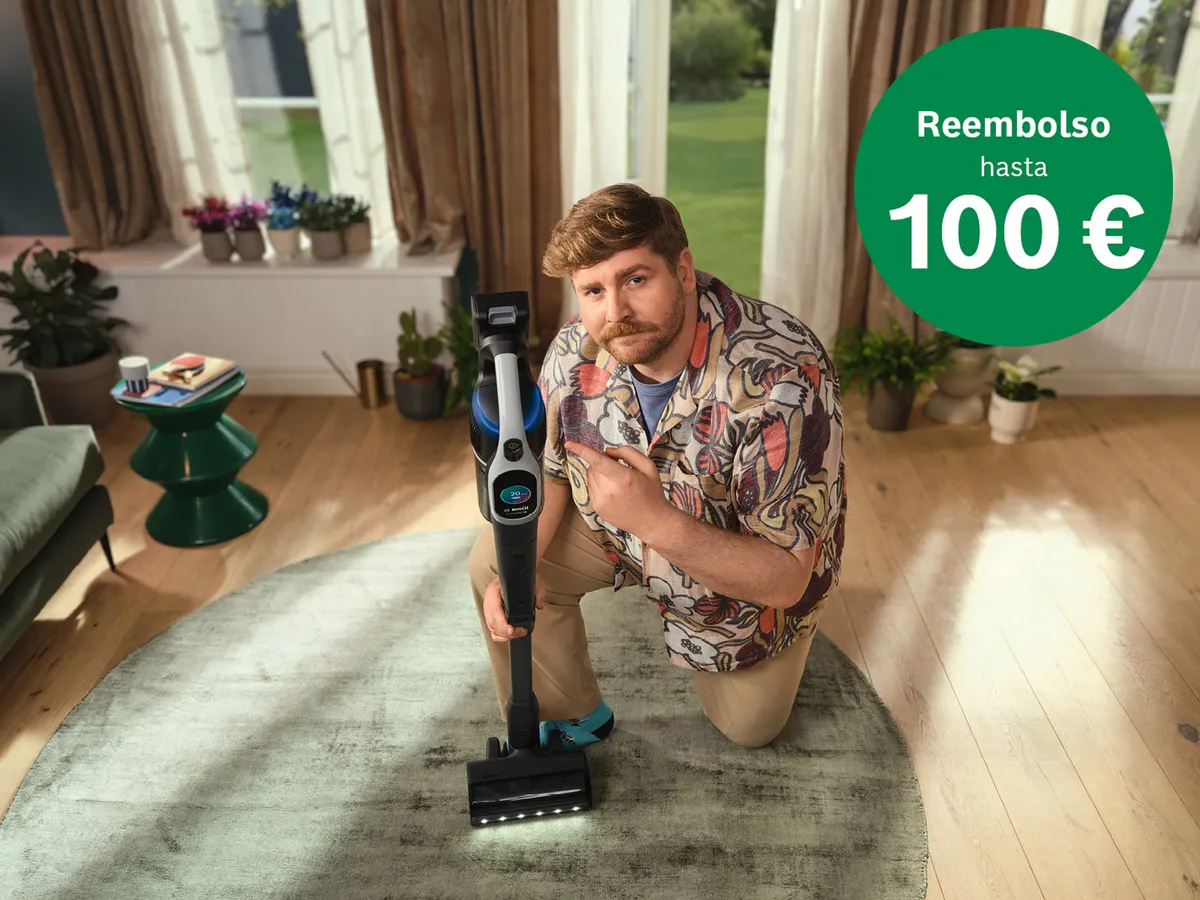 Una persona arrodillada en el suelo está mostrando una aspiradora en un ambiente luminoso y acogedor. En la esquina, hay un distintivo verde que indica "Reembolso hasta 100 €".