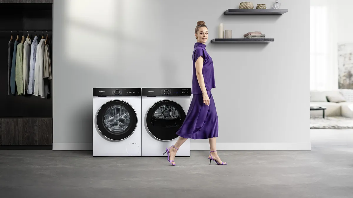 Eine Person im lässigen Outfit hockt mit ordentlich gefalteten Handtüchern neben modernen Waschmaschinen in einer stilvollen Waschküche.