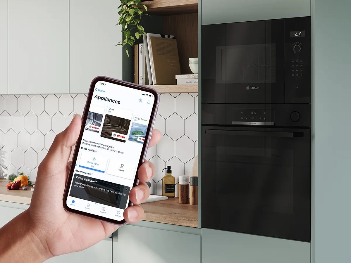 Smartphone showing the Home Connect app cookery book in front of a Bosch steam oven, highlighting smart connectivity.