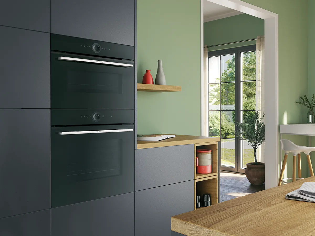 Bosch kitchen with stacked black ovens built into white cabinetry, next to a window.