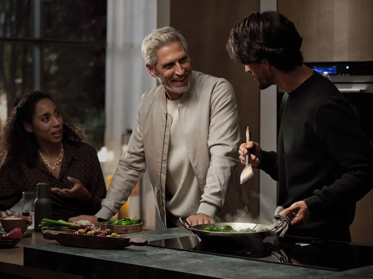 People stood around a man cooking on hob with a pan