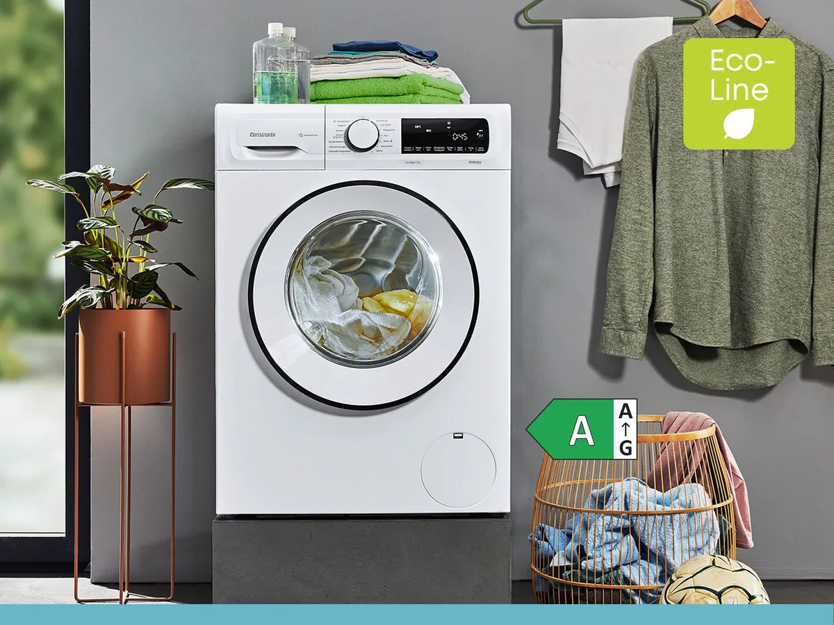 Eine Frontlader-Waschmaschine mit Wäsche steht neben einem Wäschekorb, einer Topfpflanze und einem hängenden Hemd in einem modernen Waschraum. Die Etiketten „Eco-Line“ und „Energieeffizienzklasse A“ sind sichtbar.