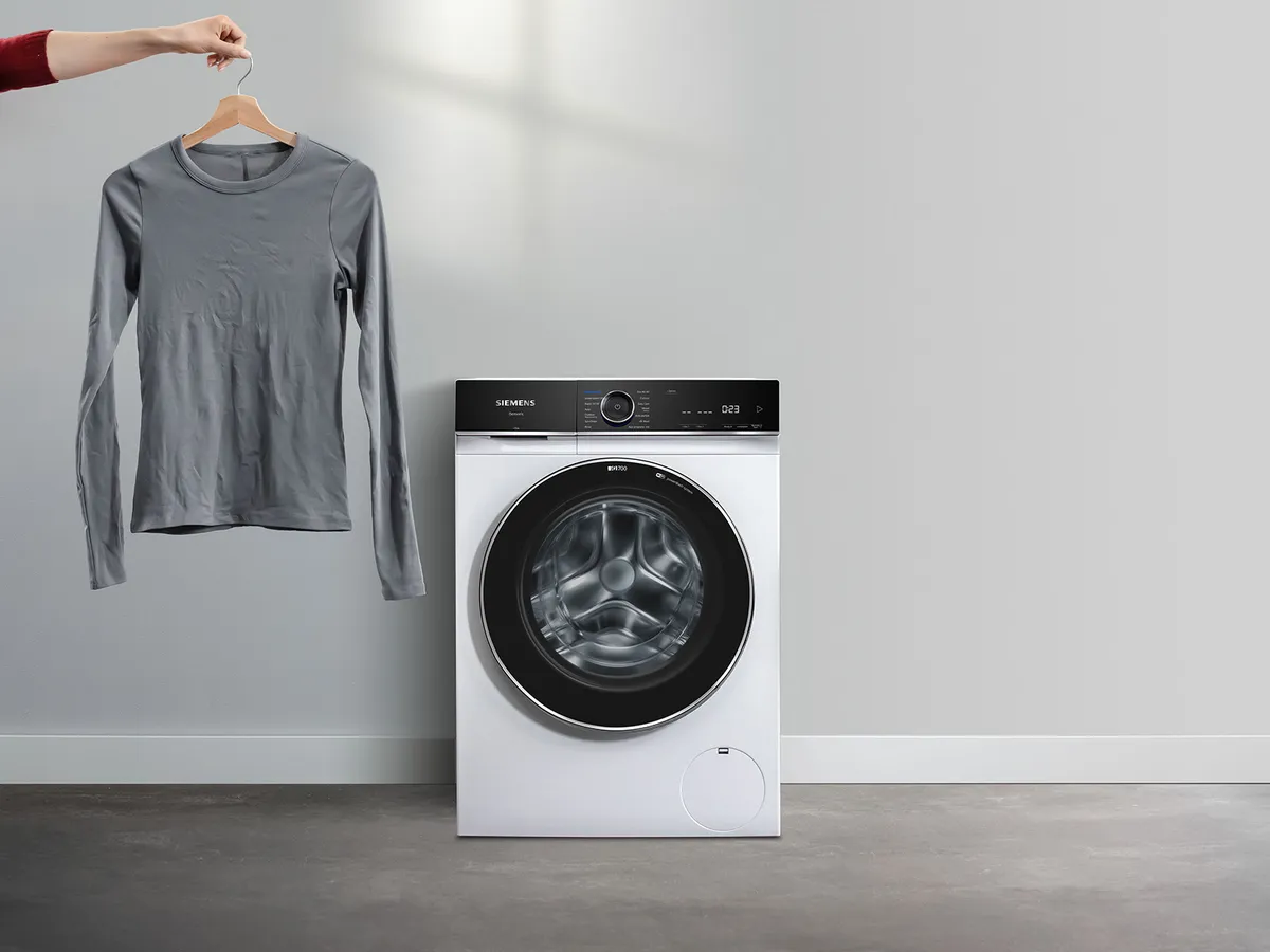 Die Hand einer Person hält ein graues Langarmhemd über einer eleganten, modernen Siemens-Waschmaschine in einem minimalistischen Waschraum.