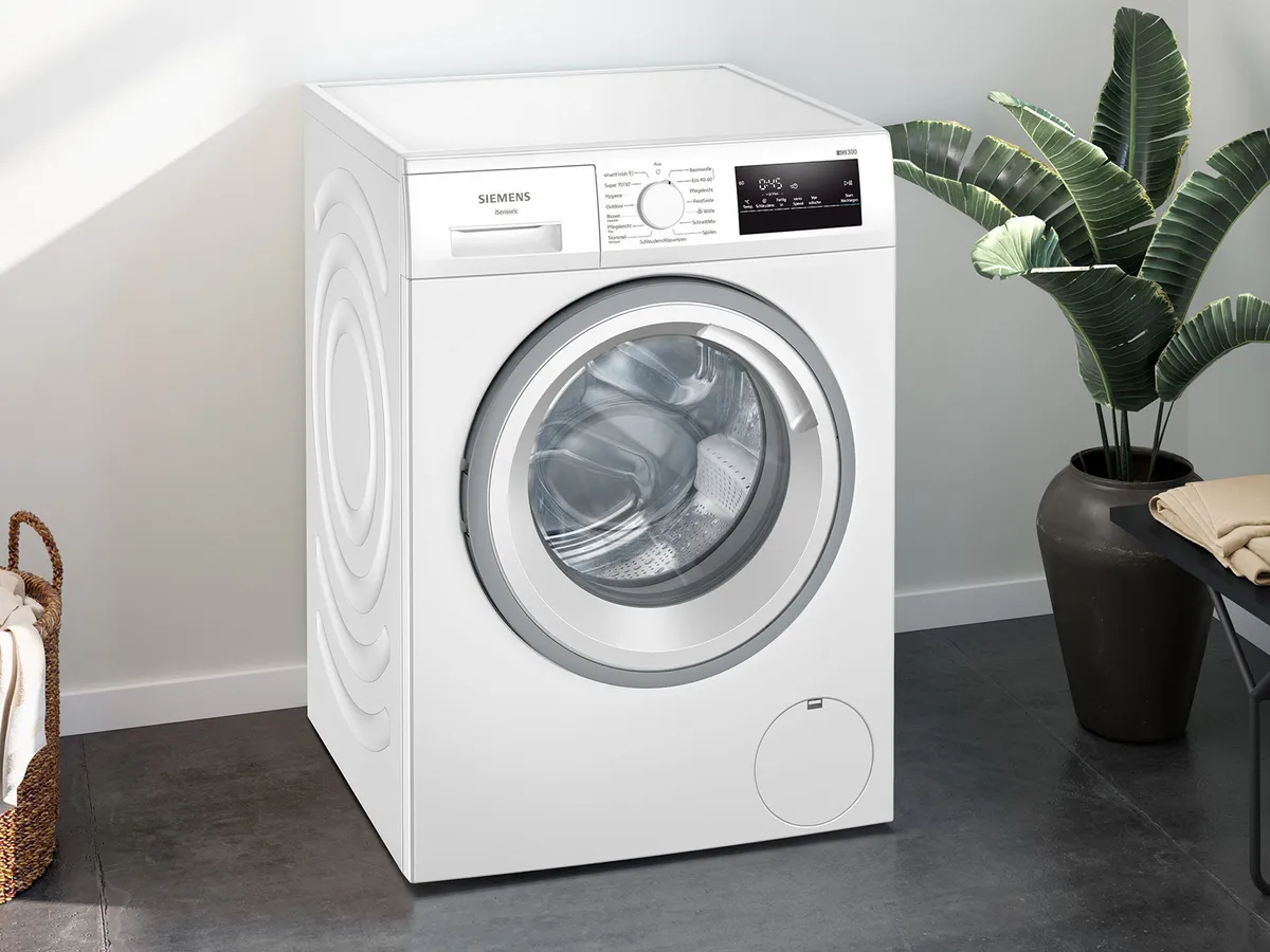 Eine elegante weiße Siemens-Waschmaschine steht an einer hellen Wand neben einer Topfpflanze und in der Nähe steht ein Wäschekorb.