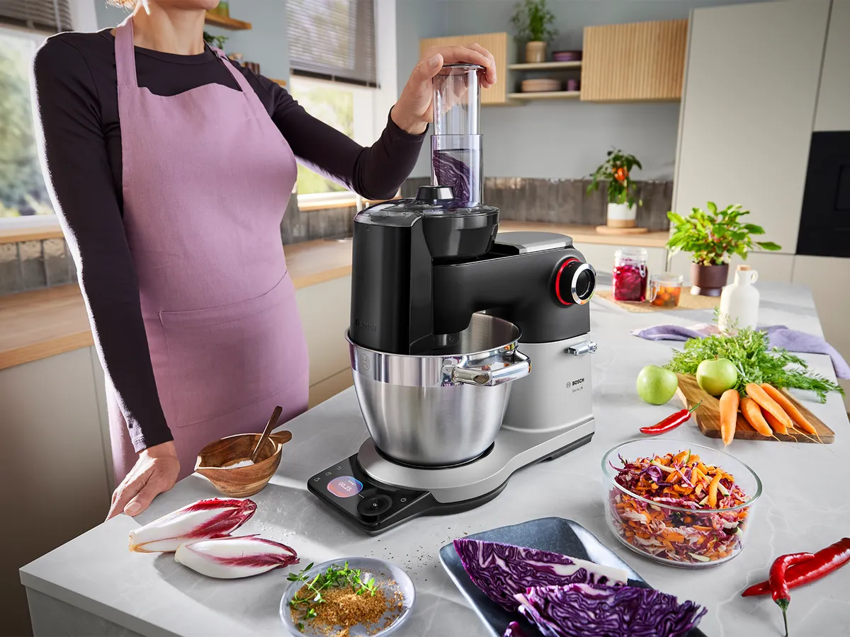 Die Serie 8 Küchenmaschine steht auf einer Küchenarbeitsplatte, umgeben von verschiedenen geschnittenen Gemüse- und Obststücken, während eine Person das Raspelzubehör verwendet.