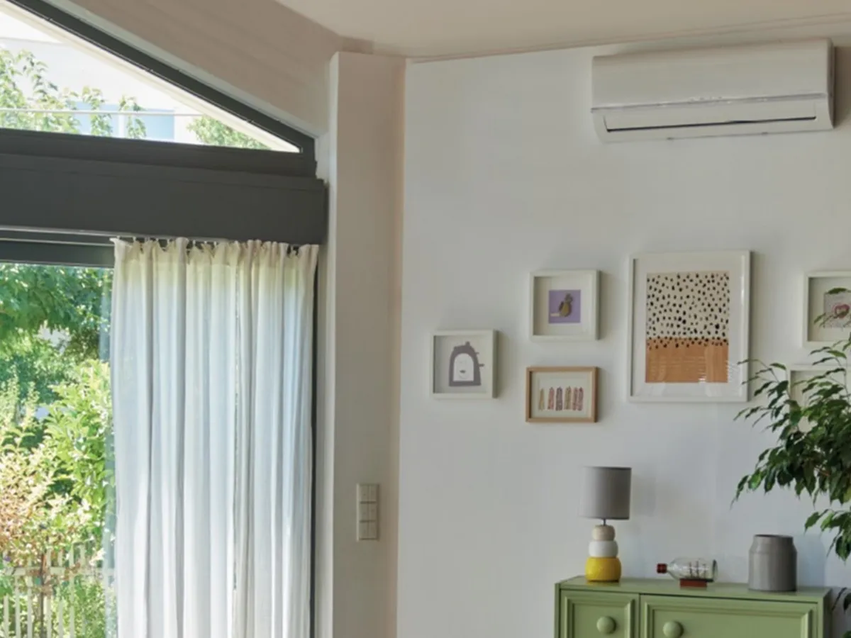 Ένα air condition με πίνακες από κατω, πράσινα φυτά στον χώρο και αριστερά μια μεγάλη μπαλκονόπορτα πυ βωλέπει στον κήπο.
