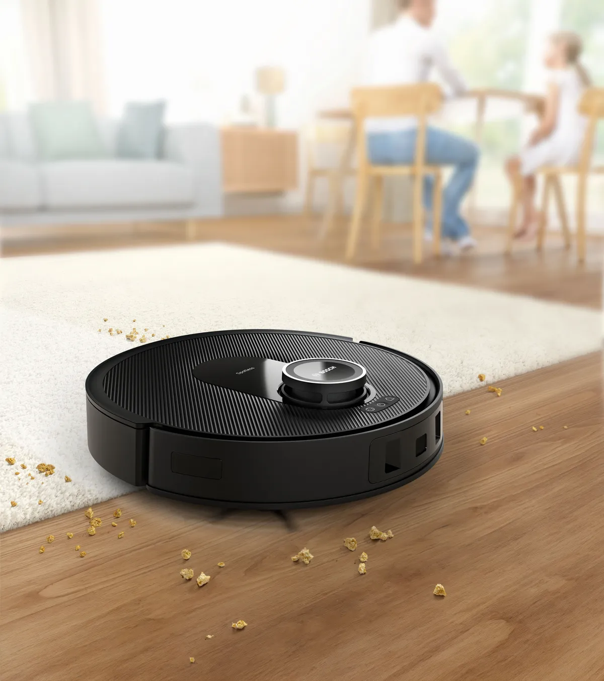 Un robot aspirapolvere nero su un pavimento di legno, vicino a briciole sparse, con una famiglia seduta a un tavolo da pranzo sullo sfondo.