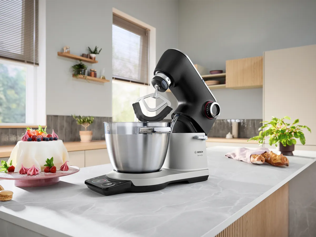Bosch kitchen mixer on a countertop with a silver bowl, cake, and plants in a modern kitchen.