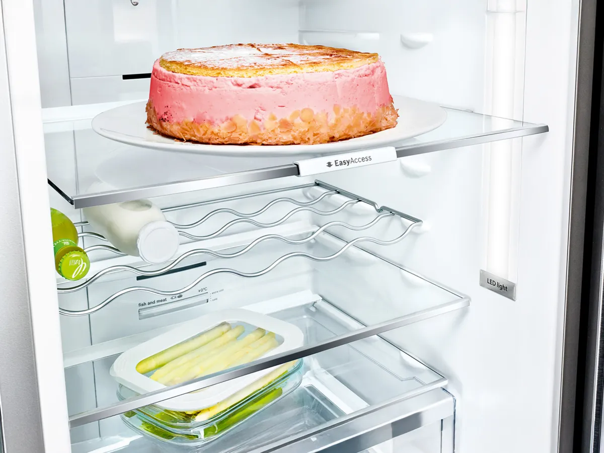 Un gâteau glacé rose posé sur une assiette blanche dans un réfrigérateur lumineux, avec des citrons verts et un récipient de céleri sur les étagères inférieures.