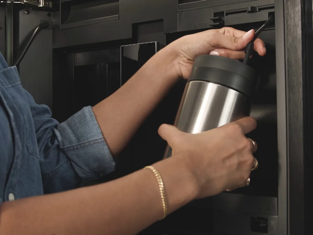 Nahaufnahme des Milchsystems eines NEFF Einbau-Kaffeevollautomaten. Eine Person verbindet den Flex-Cappuccinatore-Schlauch mit einem Edelstahl-Milchbehälter, um cremigen Milchschaum zuzubereiten.
