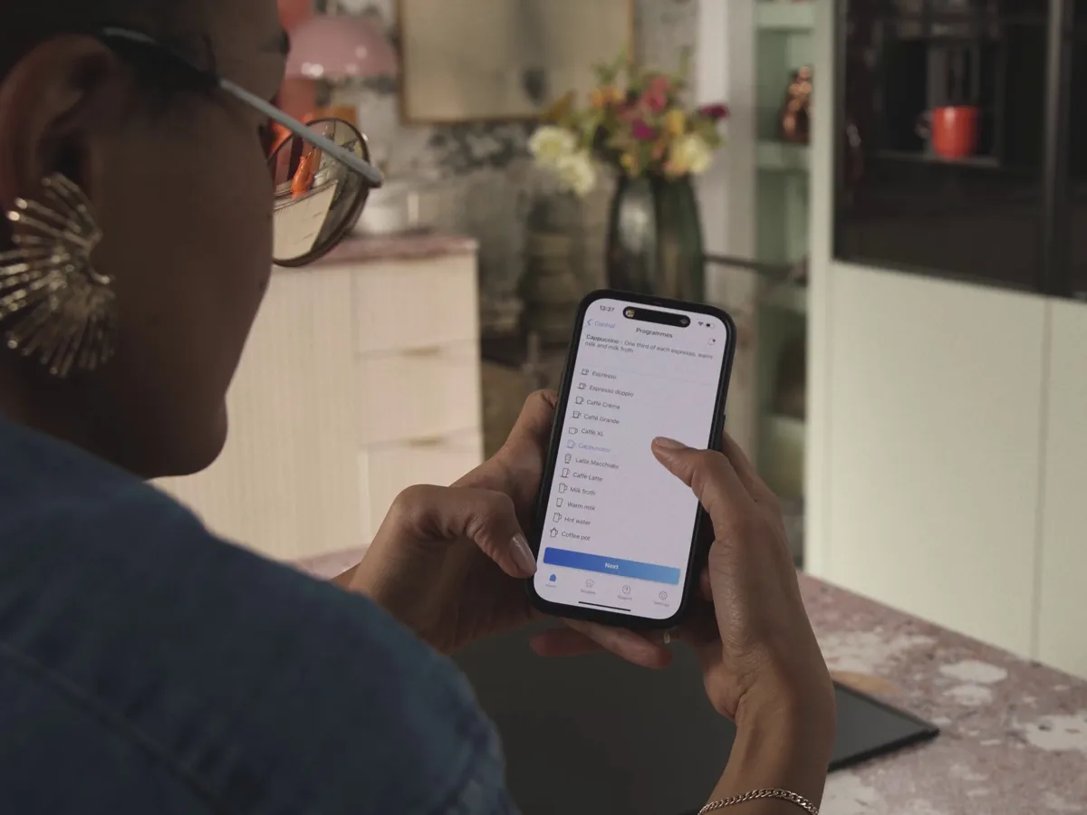 Person nutzt die Home Connect App, um Kaffeespezialitäten für den NEFF Einbau-Kaffeevollautomaten auszuwählen. Auf dem Smartphone sind verschiedene Getränke wie Espresso, Cappuccino und Latte Macchiato zu sehen.