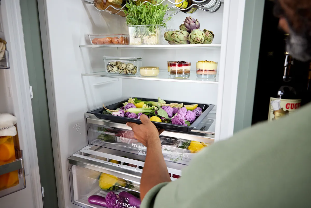 Person schiebt ein Backblech mit lila Blumenkohl und anderem Gemüse in eine Kühlschrankschublade. Im Innenraum sind oben frische Kräuter, Artischocken, Glasbehälter mit Wachteleiern und Dessertgläser zu sehen; an der Tür befinden sich Flaschen.