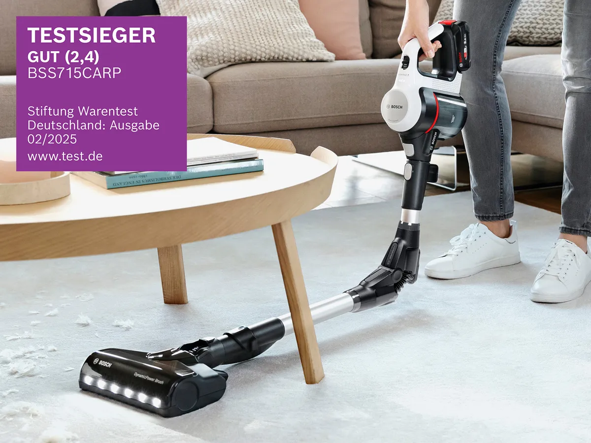 Eine Person saugt mit einem Bosch Staubsauger unter einem Couchtisch. Hinten liegt ein Kissen, auf dem Tisch liegen Bücher.