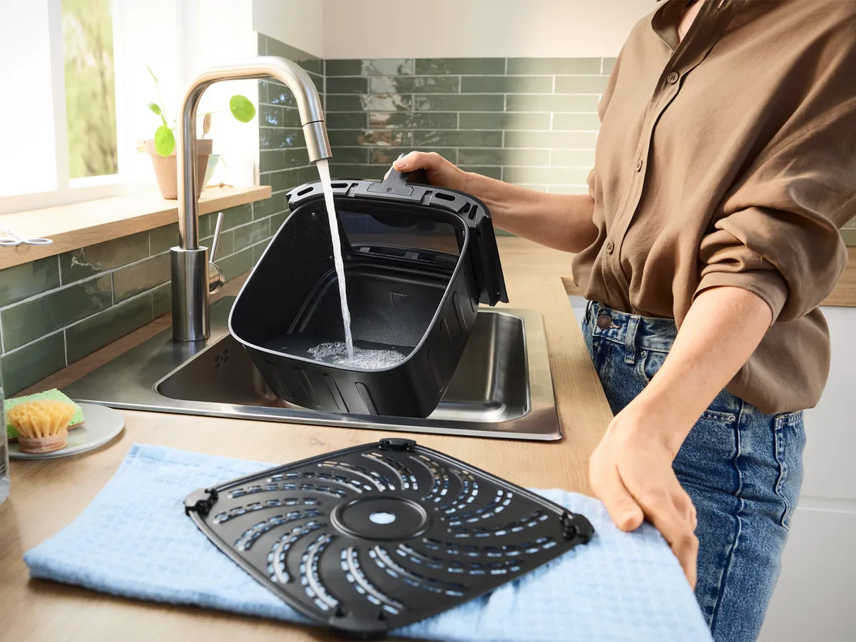 Majka ispire ladicu Bosch friteze na zrak pod tekućom vodom u kuhinjskom sudoperu, čisteći nakon kuhanja.