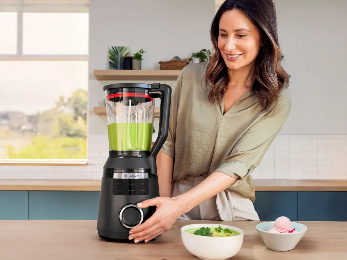 Žena se smiješi u modernoj kuhinji dok upotrebljava Bosch blender. Svježi sastojci nalaze se u zdjeli pored nje.