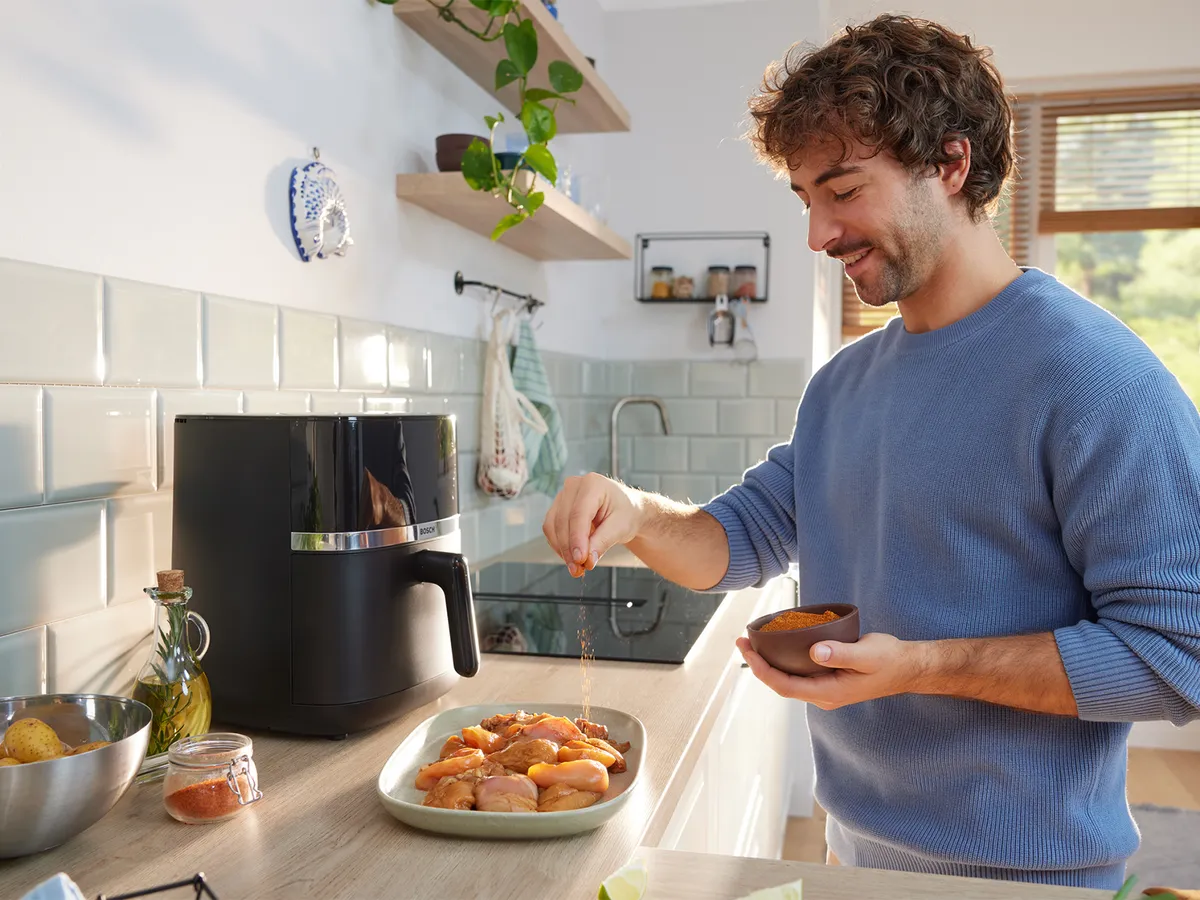 Čovjek u svijetloj kuhinji drži zdjelicu začina, posipa mješavinu začina po sirovom mesu na pultu. Bosch Air Fryer serije 4 nalazi se na pultu u pozadini.