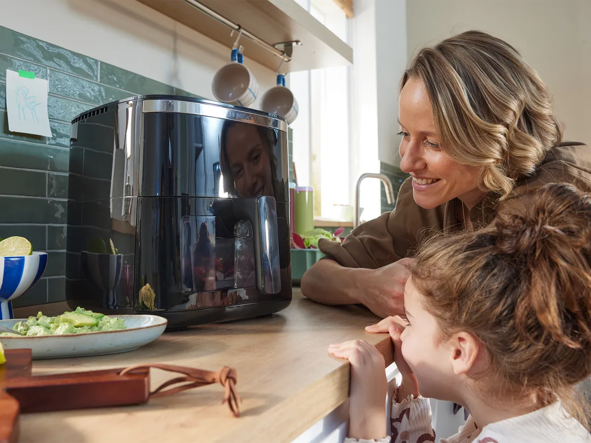Majka i kći s iščekivanjem promatraju kroz prozor Bosch Air Fryera serije 6 kako se priprema jelo. Kuhinja je svijetla i moderna.